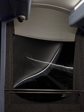 A dark tunnel view captured from the rear window of a vehicle, with strong reflections and light streaks creating an abstract effect. The window frame has a curved, futuristic design with a security camera visible on the top left corner.