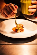 A hand carefully placing a piece of food onto a white plate using a spoon or utensil. The food appears to be artistically arranged with vibrant colors, accompanied by a sauce or puree. Another hand holds a container, possibly filled with a green sauce, with the scene illuminated warmly, emphasizing the culinary preparation.