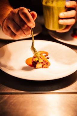 A hand carefully placing a piece of food onto a white plate using a spoon or utensil. The food appears to be artistically arranged with vibrant colors, accompanied by a sauce or puree. Another hand holds a container, possibly filled with a green sauce, with the scene illuminated warmly, emphasizing the culinary preparation.