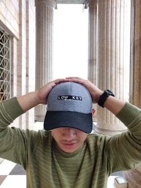 A person wearing a gray and black baseball cap with the words 'Low Key' is standing with their hands resting on top of their head. The background features tall, classical columns and architectural details, suggesting an indoor or semi-outdoor setting with stone walls.