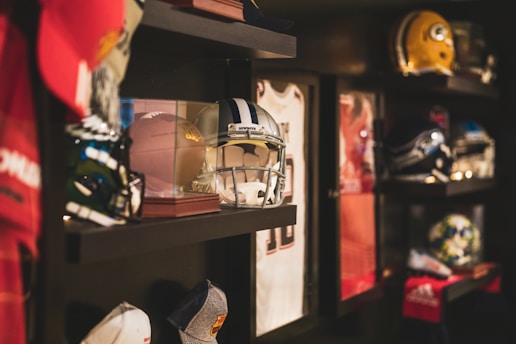 A collection of sports memorabilia displayed on shelves. Various helmets, including one with 'Cowboys' written on it, are showcased alongside a football encased in glass and framed sports jerseys. The setting is dimly lit, highlighting the items on the shelves.