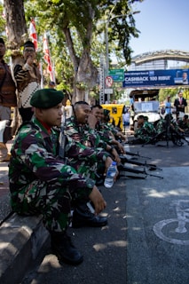 A group of uniformed soldiers in camouflage attire are seated on a curb holding rifles, with some interacting and smiling. A man stands nearby with flags in the background and a street sign visible. The scene seems to be part of a public event or gathering.