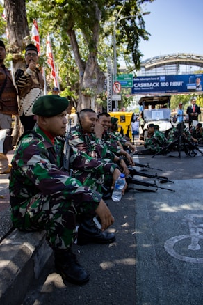 A group of uniformed soldiers in camouflage attire are seated on a curb holding rifles, with some interacting and smiling. A man stands nearby with flags in the background and a street sign visible. The scene seems to be part of a public event or gathering.