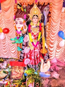 A colorful and ornate statue of a Hindu goddess is adorned with intricate jewelry and a vibrant garment. The deity has a serene expression and is surrounded by decorative elements, including balloons and garlands. Various offerings and religious items are placed in front of the statue.