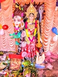 A colorful and ornate statue of a Hindu goddess is adorned with intricate jewelry and a vibrant garment. The deity has a serene expression and is surrounded by decorative elements, including balloons and garlands. Various offerings and religious items are placed in front of the statue.