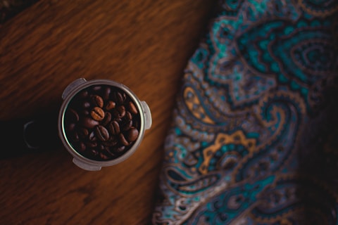 A portafilter filled with dark roasted coffee beans sits on a wooden surface next to a colorful, paisley-patterned fabric. The fabric features swirling designs in shades of blue, gold, and brown. The scene is dimly lit, creating a cozy and inviting atmosphere.