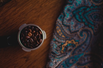 A portafilter filled with dark roasted coffee beans sits on a wooden surface next to a colorful, paisley-patterned fabric. The fabric features swirling designs in shades of blue, gold, and brown. The scene is dimly lit, creating a cozy and inviting atmosphere.
