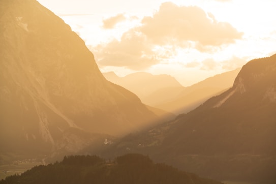 Warm, golden light bathes a mountainous landscape with layers of rugged peaks and valleys. The sunlight streams through and casts long shadows across the scene, while fluffy clouds hover in the sky. The atmosphere is calm and serene, with a hint of a small settlement visible in the valley below.