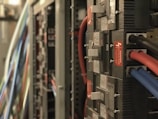 A close-up view of a server room with multiple electrical panels and cables. Several red, blue, and black cables are connected to the units, which have labels and warning signs indicating danger.