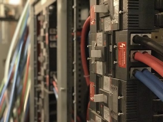 A close-up view of a server room with multiple electrical panels and cables. Several red, blue, and black cables are connected to the units, which have labels and warning signs indicating danger.