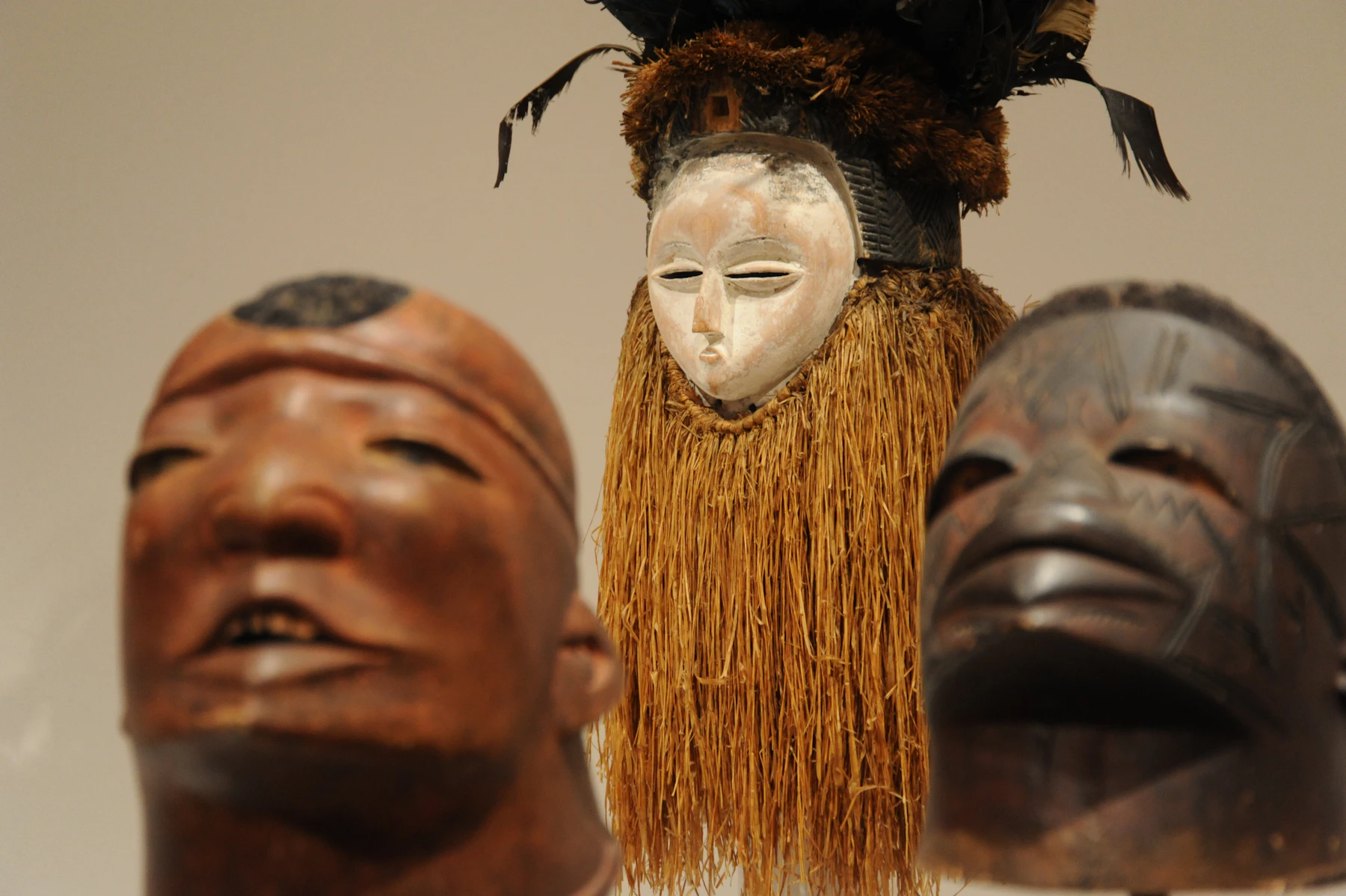 A white-faced mask with a long raffia beard between two dark carved wooden masks in a museum case