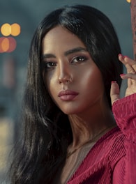 A woman with long, dark hair and a nose ring gazes directly into the camera. She is wearing a red, textured blouse and has her hand touching a vertical surface. Soft lighting enhances her features, creating a serene and introspective mood.
