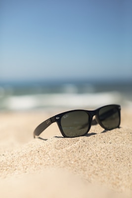 A sleek pair of sunglasses resting on a sandy beach towel with a hint of ocean waves in the background.
