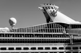 A sidelong view of a large cruise ship with a prominent glass-covered upper deck and a distinctive logo on the smokestack. The ship's design incorporates a variety of geometric shapes, with a sphere-like structure also visible on the deck.