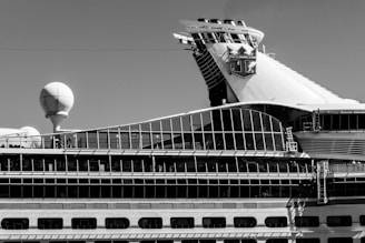 A sidelong view of a large cruise ship with a prominent glass-covered upper deck and a distinctive logo on the smokestack. The ship's design incorporates a variety of geometric shapes, with a sphere-like structure also visible on the deck.