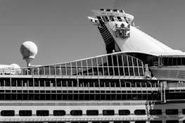 A sidelong view of a large cruise ship with a prominent glass-covered upper deck and a distinctive logo on the smokestack. The ship's design incorporates a variety of geometric shapes, with a sphere-like structure also visible on the deck.
