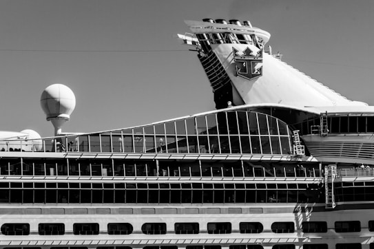 A sidelong view of a large cruise ship with a prominent glass-covered upper deck and a distinctive logo on the smokestack. The ship's design incorporates a variety of geometric shapes, with a sphere-like structure also visible on the deck.