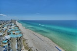 An aerial view of a long, sandy beach stretching along the coast with turquoise waters extending to the horizon. Modern multi-story buildings line the shoreline, and rows of blue umbrellas are neatly arranged on the beach. The sky is clear with only a few clouds visible, indicating sunny weather.