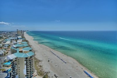 An aerial view of a long, sandy beach stretching along the coast with turquoise waters extending to the horizon. Modern multi-story buildings line the shoreline, and rows of blue umbrellas are neatly arranged on the beach. The sky is clear with only a few clouds visible, indicating sunny weather.