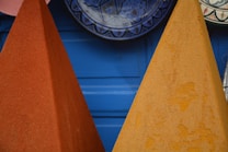 A close-up of two conical piles of colorful spices or powders in bright orange and yellow hues against a blue wooden panel background. Decorative plates with intricate designs are partially visible hanging above.