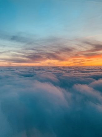 A stunning aerial view of fluffy clouds with a vibrant sunset in the background. The sky transitions from deep blue to warm orange, creating a serene and peaceful atmosphere. The clouds appear soft and dense, adding depth to the scene.