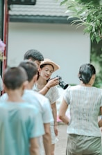 A group of people gather outdoors for a photography session. A woman in a straw hat holds a camera and focuses on taking a picture. Several people stand around her, casually dressed, and the scene has a relaxed, candid atmosphere.