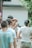 A group of people gather outdoors for a photography session. A woman in a straw hat holds a camera and focuses on taking a picture. Several people stand around her, casually dressed, and the scene has a relaxed, candid atmosphere.