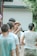 A group of people gather outdoors for a photography session. A woman in a straw hat holds a camera and focuses on taking a picture. Several people stand around her, casually dressed, and the scene has a relaxed, candid atmosphere.