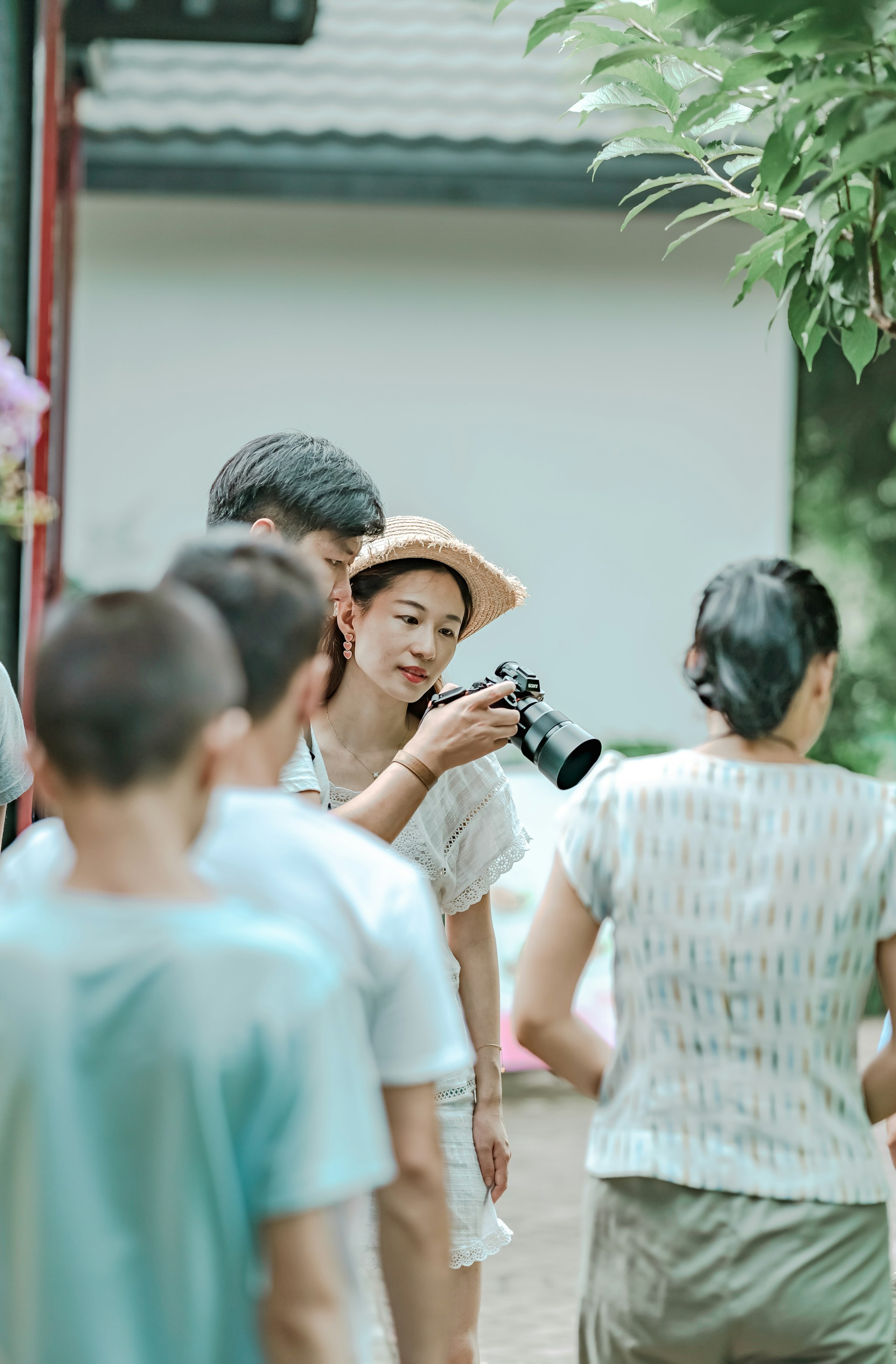 A group of people gather outdoors for a photography session. A woman in a straw hat holds a camera and focuses on taking a picture. Several people stand around her, casually dressed, and the scene has a relaxed, candid atmosphere.