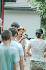 A group of people gather outdoors for a photography session. A woman in a straw hat holds a camera and focuses on taking a picture. Several people stand around her, casually dressed, and the scene has a relaxed, candid atmosphere.