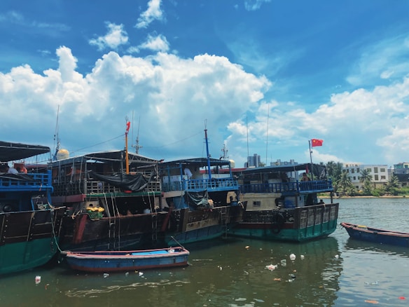 Several large boats are docked near the shoreline, with buildings and trees visible in the background. The sky is partly cloudy, creating a bright and inviting atmosphere. Flags are displayed on some of the boats, and small items or debris are floating in the water alongside them.