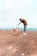 A person wearing a brown shirt, jeans, and a cap bends over to offer a treat to a tan dog on a rocky terrain. The background features a wide expanse of blue ocean under a pale sky.