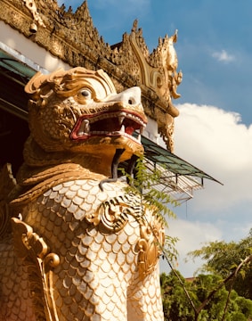 A large, intricately designed statue of a mythical creature with scales and a fierce expression, featuring ornate gold detailing against a backdrop of blue sky and clouds. The statue is positioned near architectural elements that also have elaborate decorations, and there's greenery in the foreground.