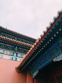 The image features the intricately decorated roof of a traditional building with colorful patterns and detailed craftsmanship. The roof tiles are arranged symmetrically, and artistic designs are visible in vibrant colors like blue, red, and gold. The background is a pale, cloudy sky that contrasts with the bold architecture.