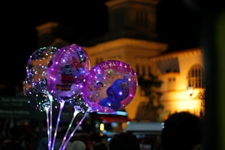 Several transparent balloons illuminated with colorful LED lights float against a nighttime city backdrop. The balloons feature cartoon characters and emit a magical, festive glow. The out-of-focus background reveals softly lit buildings with a classic architectural style.