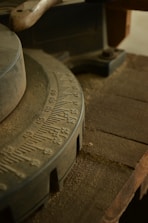 A close-up view of a worn, industrial piece of machinery with a dusty surface. The focus is on a circular gauge with numerals etched into its metal surface, likely part of a larger tool or machine. The surrounding area includes rough, wooden elements and a handle suggesting usage in a workshop setting.