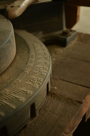 A close-up view of a worn, industrial piece of machinery with a dusty surface. The focus is on a circular gauge with numerals etched into its metal surface, likely part of a larger tool or machine. The surrounding area includes rough, wooden elements and a handle suggesting usage in a workshop setting.