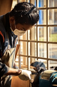 A person wearing a face mask and gloves is working with a bench grinder near a window with sunlight streaming through. The atmosphere suggests a workshop setting.