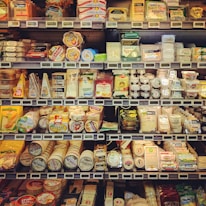 A well-stocked supermarket dairy section featuring a wide variety of cheeses displayed on shelves. The assortment includes round, rectangular, and wedge-shaped packages with labels in multiple languages. Prices are visible on digital screens beneath each item.