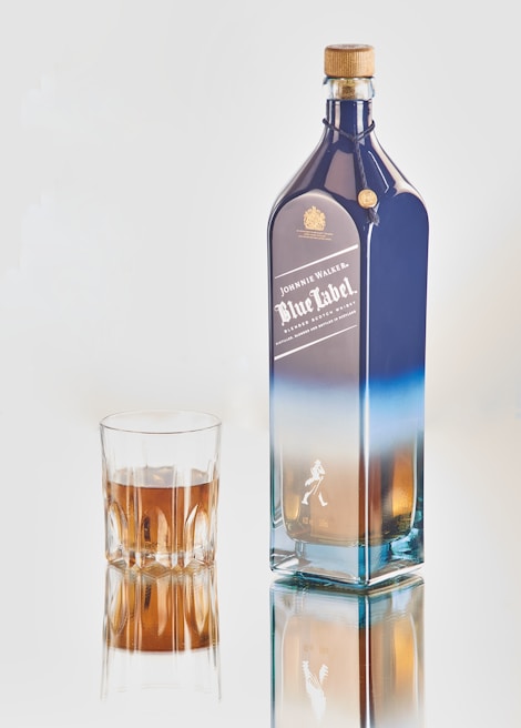 A tall, elegant bottle of whiskey with a blue label next to a glass filled with a brown liquid. The bottle features a gold cap and has branding and logos on its front.