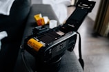 A film camera with its back open exposing a roll of 35mm film that is loaded inside. The camera is placed on a soft surface, possibly a couch, and there are film boxes nearby, suggesting a setup for photography.