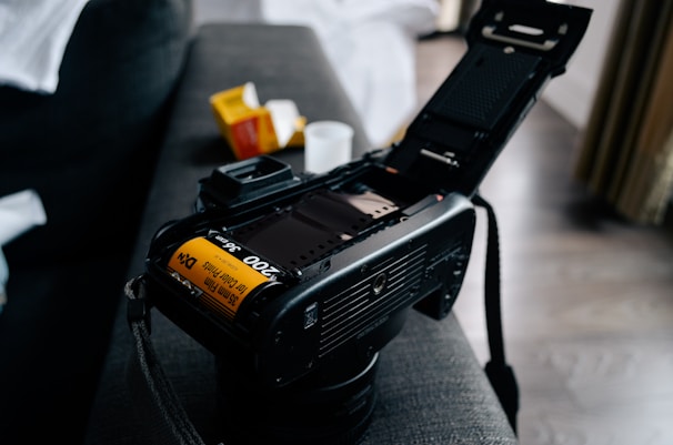 A film camera with its back open exposing a roll of 35mm film that is loaded inside. The camera is placed on a soft surface, possibly a couch, and there are film boxes nearby, suggesting a setup for photography.