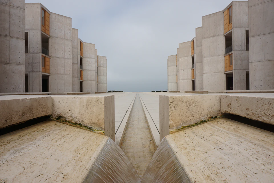 The image presents a large, rectangular concrete structure with multiple levels and balconies on each floor. The building is situated next to a body of water, which can be seen flowing through the center of the structure. The perspective of the photo is taken from below, looking up at the building's impressive height and architectural design.

The sky above the scene is overcast, casting a soft light that enhances the gray tones of the concrete structure. The image does not contain any discernible text or human figures, making it an interesting visual study in architecture and natural elements.