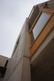 A modern architectural structure with a focus on concrete and wood materials. The angle emphasizes the geometric forms and the interaction between the sky and the building. A ladder is visible near the top, adding an industrial touch.
