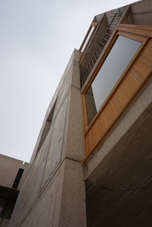 A modern architectural structure with a focus on concrete and wood materials. The angle emphasizes the geometric forms and the interaction between the sky and the building. A ladder is visible near the top, adding an industrial touch.