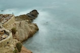 A scenic rocky coastline with a stone patio overlooking the ocean. Wooden tables and chairs are arranged along the patio, creating a dining area with a view of the sea. The ocean water appears misty and flows gently around the rocks.