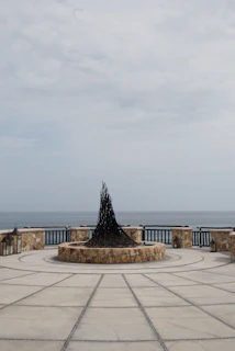 A circular stone platform by the sea features a central sculpture with a conical shape made of dark, intertwined branches or metal pieces. The platform is enclosed by a stone wall with integrated lanterns and has a geometric pattern on the ground. The ocean is visible in the background under a cloudy sky.