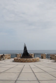 A circular stone platform by the sea features a central sculpture with a conical shape made of dark, intertwined branches or metal pieces. The platform is enclosed by a stone wall with integrated lanterns and has a geometric pattern on the ground. The ocean is visible in the background under a cloudy sky.