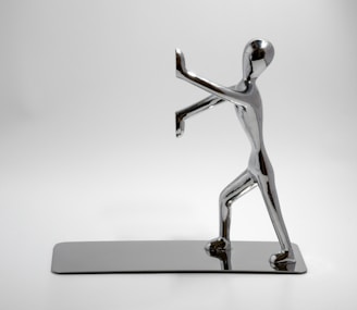 A chrome-plated sculpture of a human figure in a dynamic pose, with one foot forward and arms raised, appearing to push against an invisible barrier. The sculpture is highly polished and reflective, set on a matching rectangular base, all against a plain white background.
