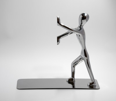 A chrome-plated sculpture of a human figure in a dynamic pose, with one foot forward and arms raised, appearing to push against an invisible barrier. The sculpture is highly polished and reflective, set on a matching rectangular base, all against a plain white background.